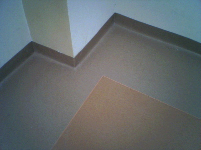 Szczegółowe ujęcie narożnika pomieszczenia z nową, jednolitą posadzką z PVC w odcieniach beżu i brązu, z widoczną listwą przypodłogową w kolorze ciemnego brązu.